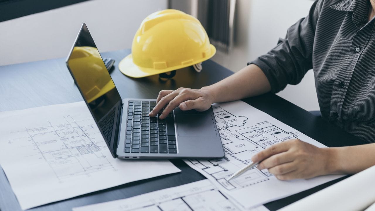 Best Architectural Planning Software: The Secret to Creative Design for Your Future Projects صورة بها جهاز لاب توب وخوذة صفراء ويد رجل يجلس على مكتب وأمامه بعض الأوراق